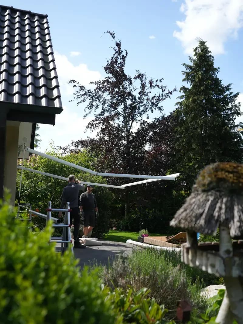 Montage von Sonnenschutz in Gütersloh: Zwei Handwerker bauen im Garten eine Markise auf, Maßanfertigung für Markisen und Rollläden in OWL.
